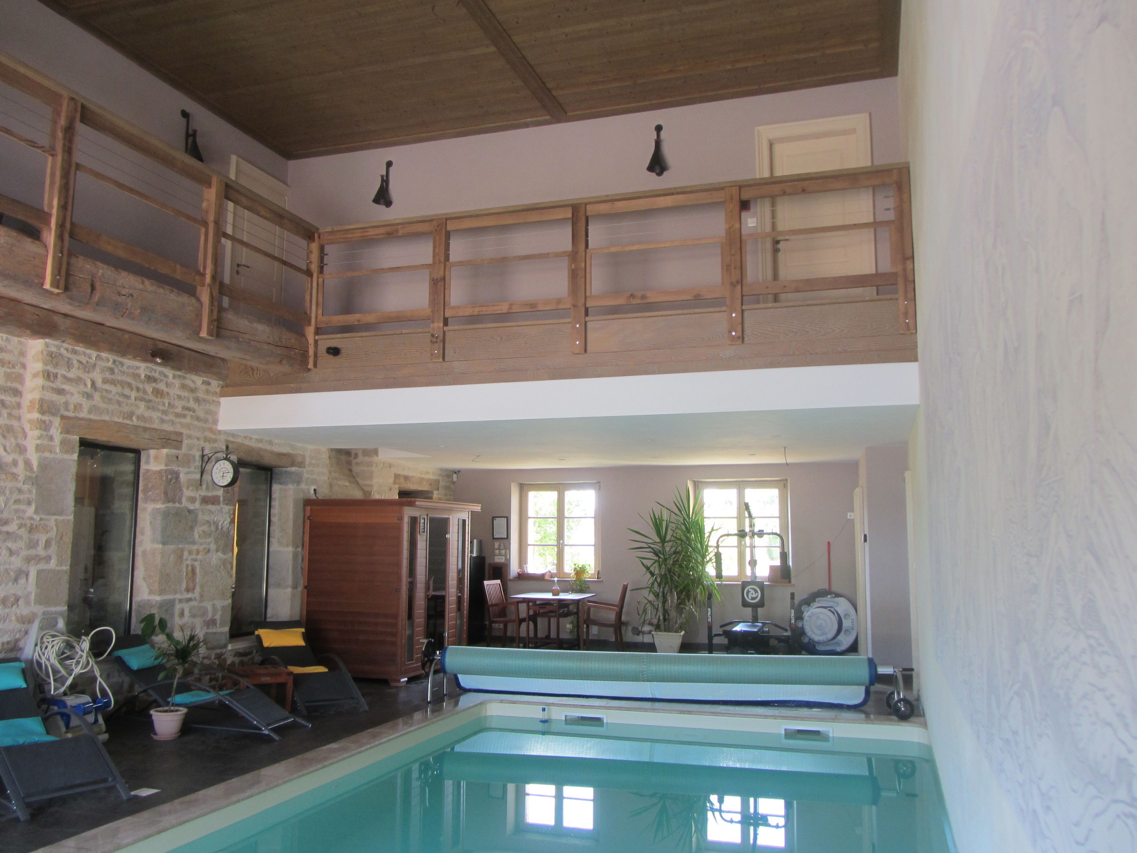 A spacious indoor swimming pool with a wooden deck above, stone walls, and a dining area with natural light.