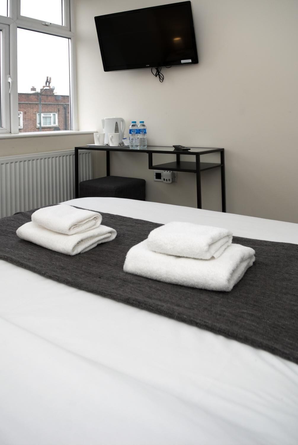 Una habitación de hotel con una cama, un televisor de pantalla plana montado en la pared, una mesita con una tetera, tazas y botellas de agua.