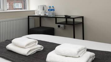 Una habitación de hotel con una cama, un televisor de pantalla plana montado en la pared, una mesita con una tetera, tazas y botellas de agua.