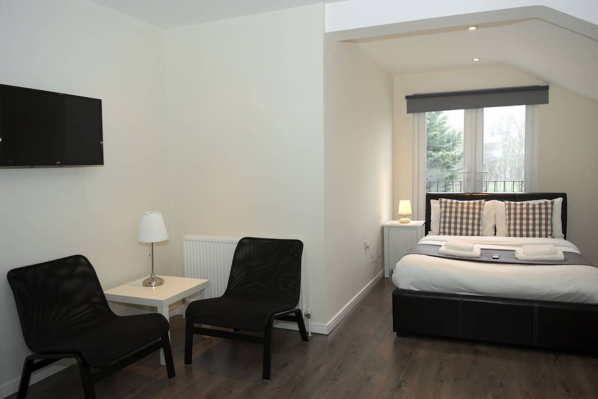 A bedroom with a bed, two chairs, a TV, a lamp, and a window.