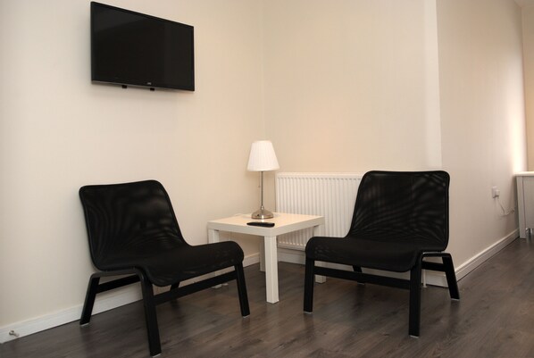 Una habitación con dos sillas negras, una mesa blanca, una lámpara y un televisor montado en la pared.
