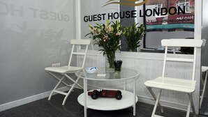 A reception area with a glass table, two white folding chairs, a vase with flowers, and a toy car on a shelf. - Guest House London (London)