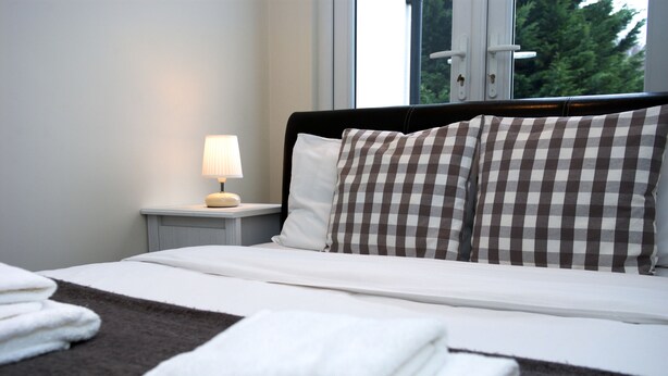 A neatly made bed with checkered pillows, a bedside lamp, and folded towels on a dark brown bedspread. - Guest House London (London)