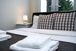 Una cama bien tendida con almohadas a cuadros, una lámpara de noche y toallas dobladas sobre un edredón marrón oscuro.