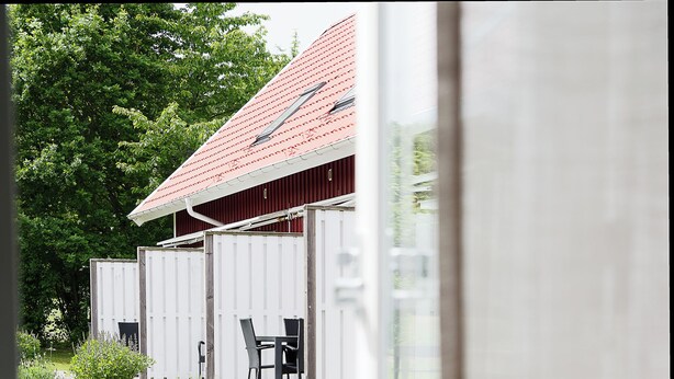 A wooden deck with a table and chairs, a covered outdoor seating area, and a view of a red-roofed building. - Apelviken Lägenhetshotell (Varberg)