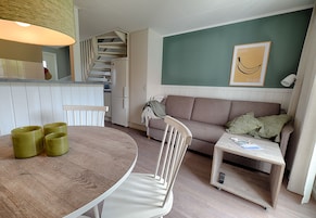 Un espacio habitable compacto con una mesa de comedor, un sofá, una mesita auxiliar y una zona de cocina con una escalera.