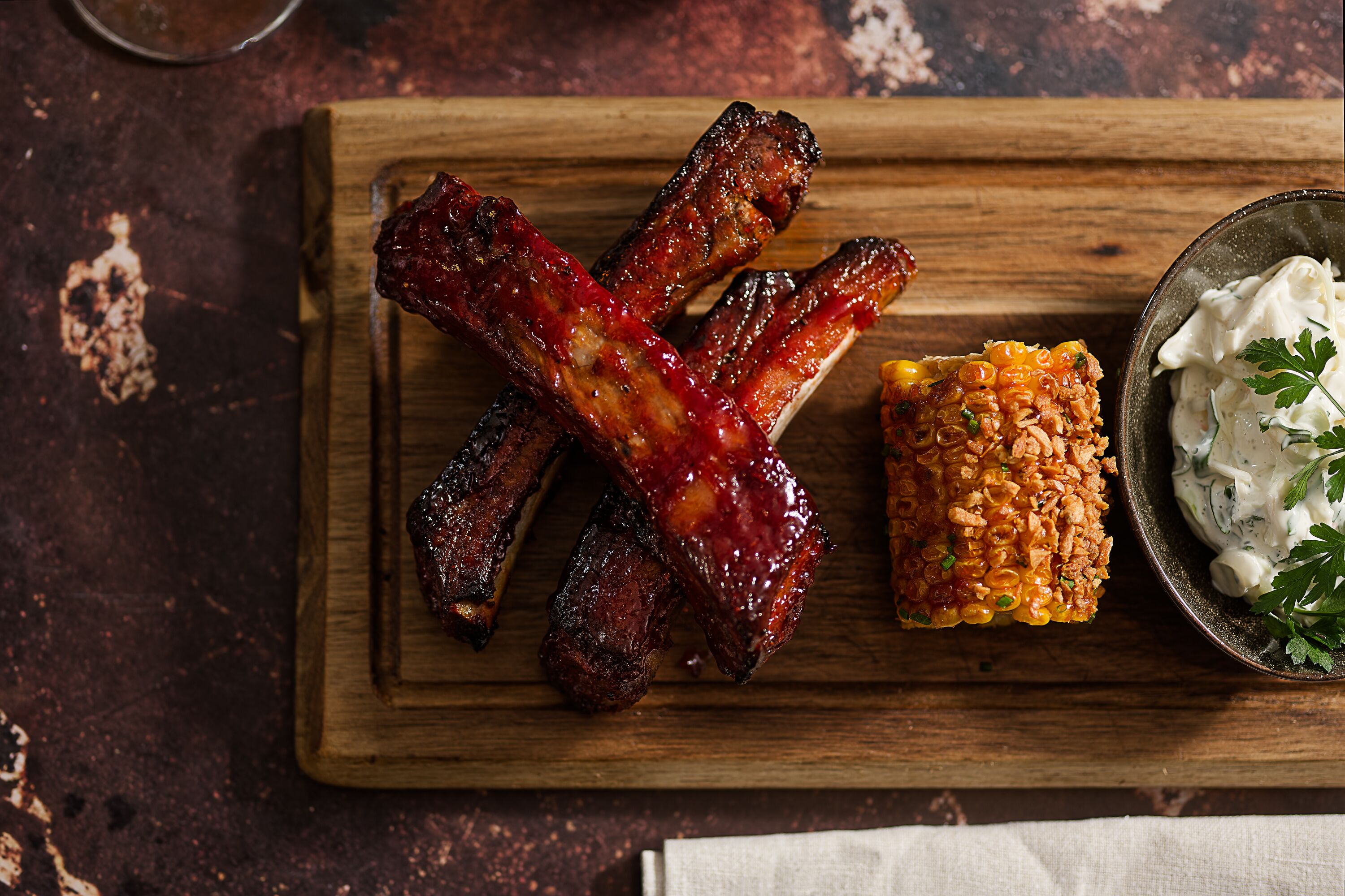A wooden cutting board with barbecued ribs and a side of corn on the cob.