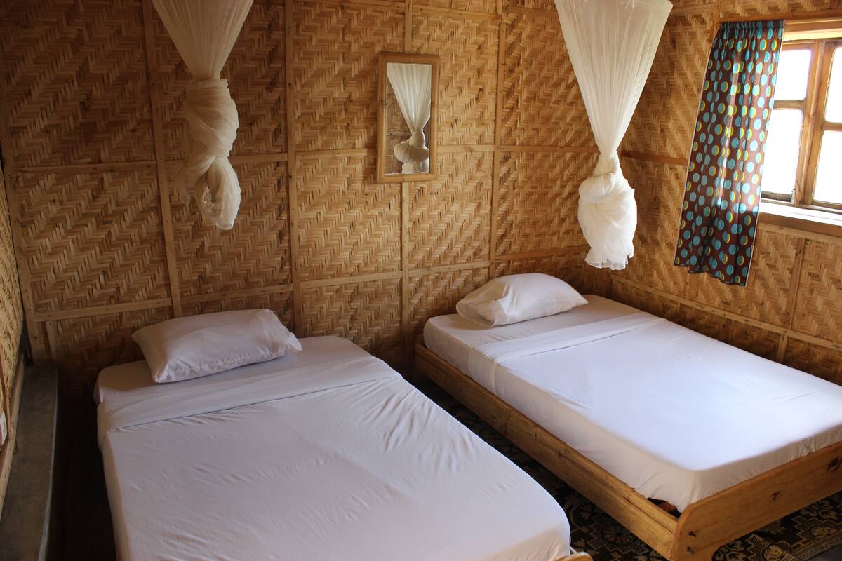 A room with two wooden beds, white bedding, a woven wall pattern, a window with curtains, and a hanging white canopy.