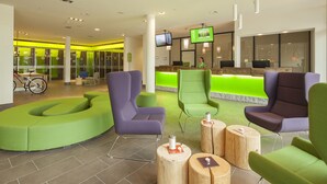 A modern reception area with green and purple seating, wooden tables, and a TV screen displaying a program. - Explorer Hotel Ötztal (Umhausen)
