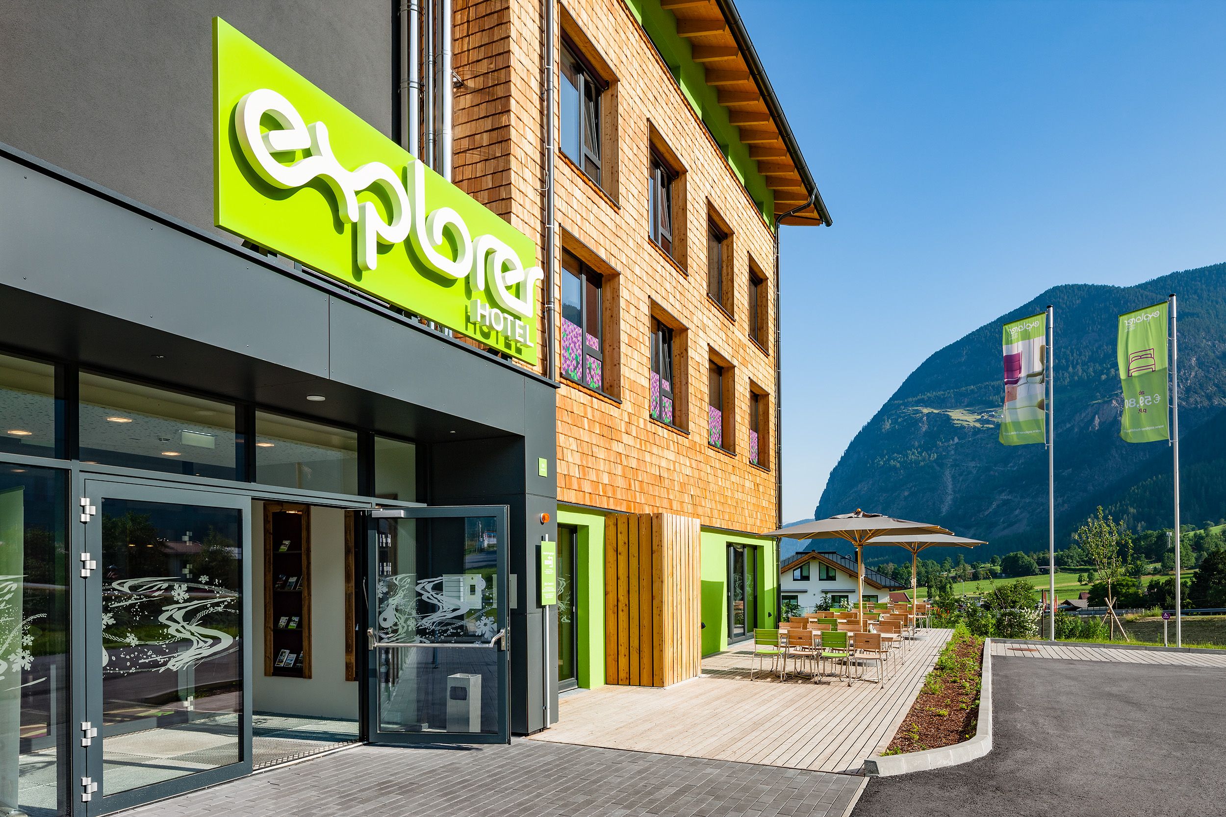 Un hotel con un llamativo letrero verde, fachada de madera y vistas a montañas al fondo.