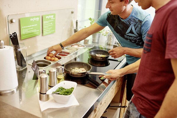 A kitchen counter with a frying pan, a bowl of green vegetables, and a container of eggs. There are two green signs on the wall, a metal container with utensils, and a white paper towel dispenser. - Explorer Hotel Ötztal (Umhausen)