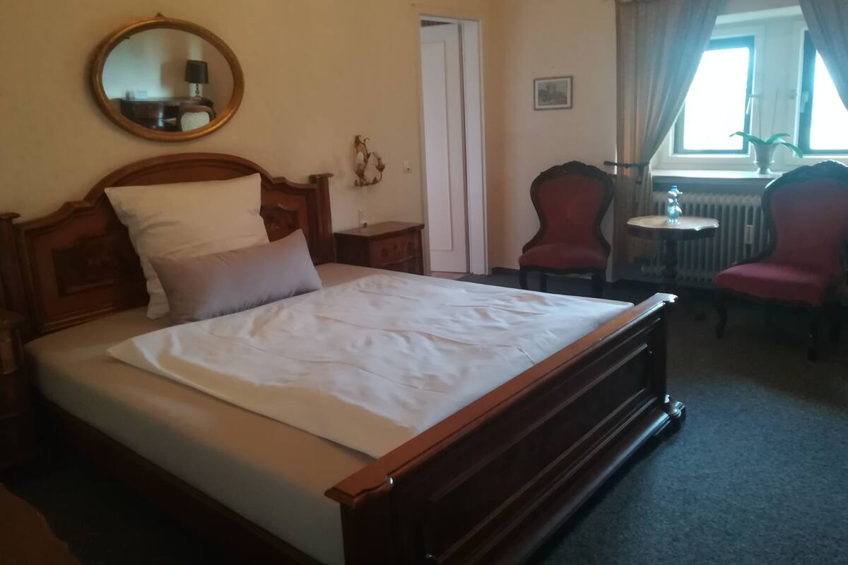 A bedroom with a wooden bed, a round mirror, a red chair, and a window with curtains.