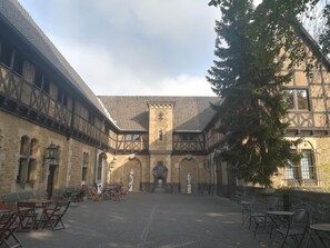 A courtyard with a building featuring a clock tower, stone walls, and traditional half-timbered architecture. Tables and chairs are arranged for outdoor seating. - Kommende Ramersdorf (Bonn)