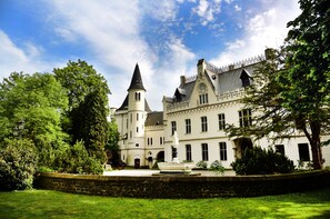 Ein großes, historisches Gebäude mit einem markanten Turm und zahlreichen Fenstern, umgeben von üppigem Grün und einer Steinmauer.