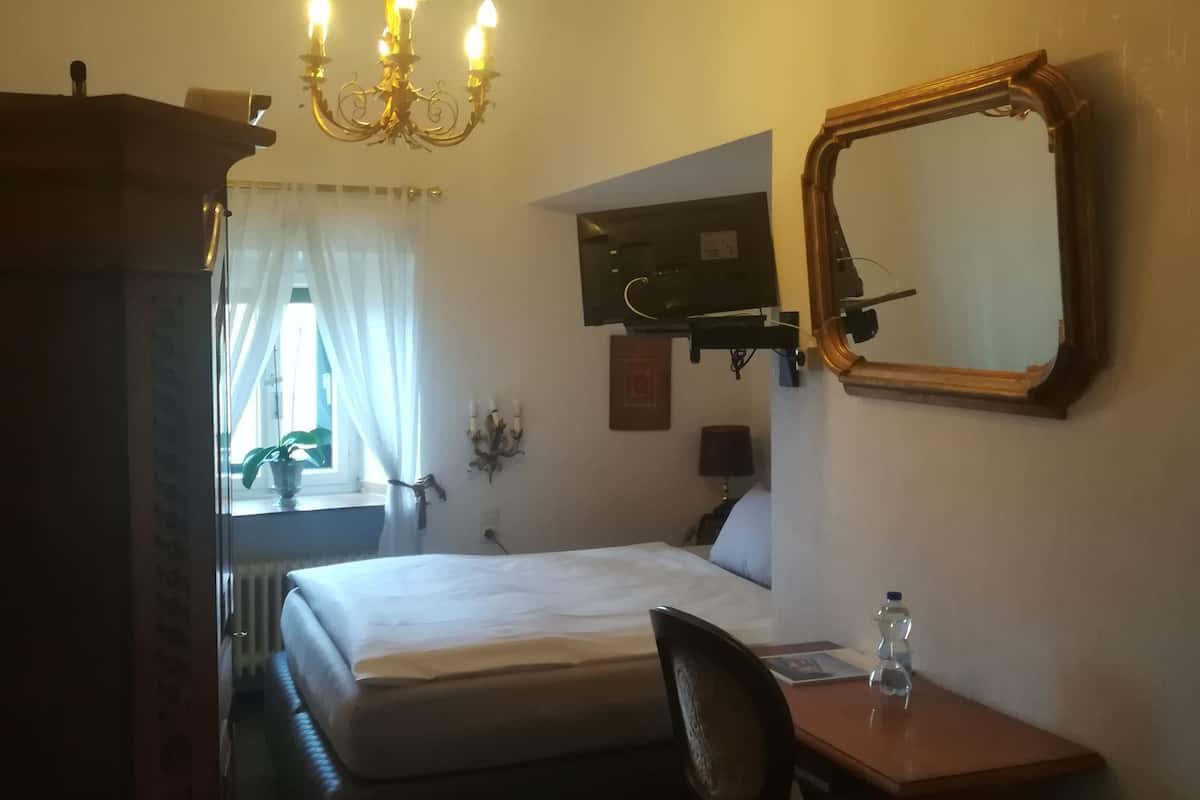 A hotel room with a bed, a desk with a chair, a mirror, a chandelier, and a window with curtains.