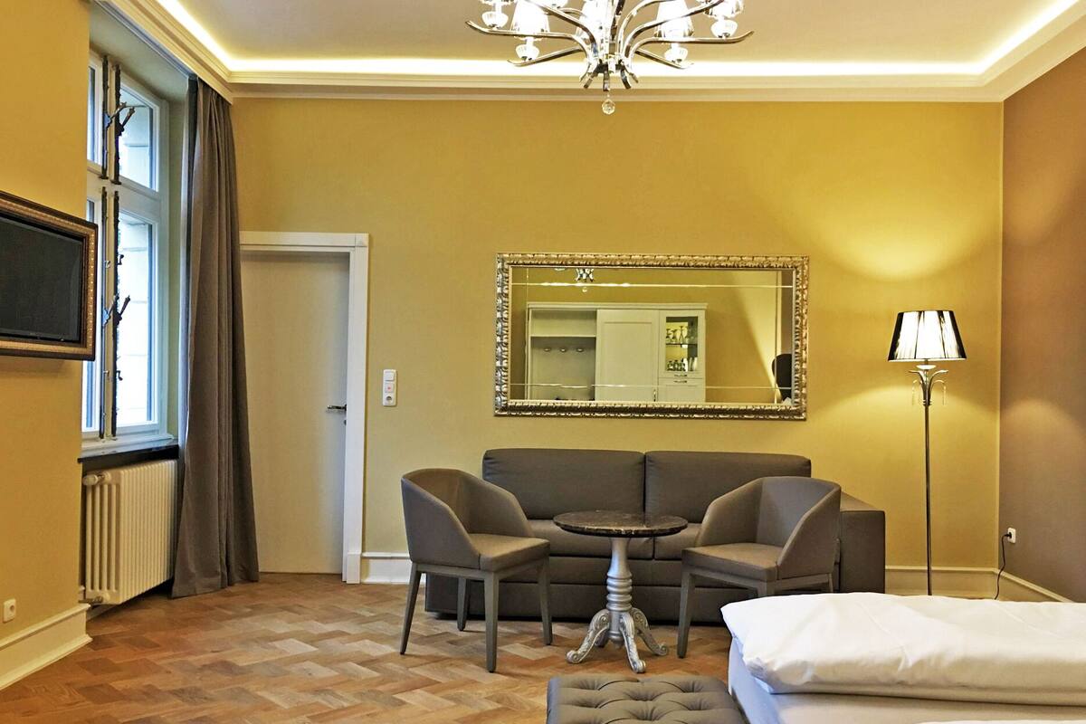 A hotel room with a sofa, two armchairs, a small round table, a bed, a large mirror, a floor lamp, and a radiator.