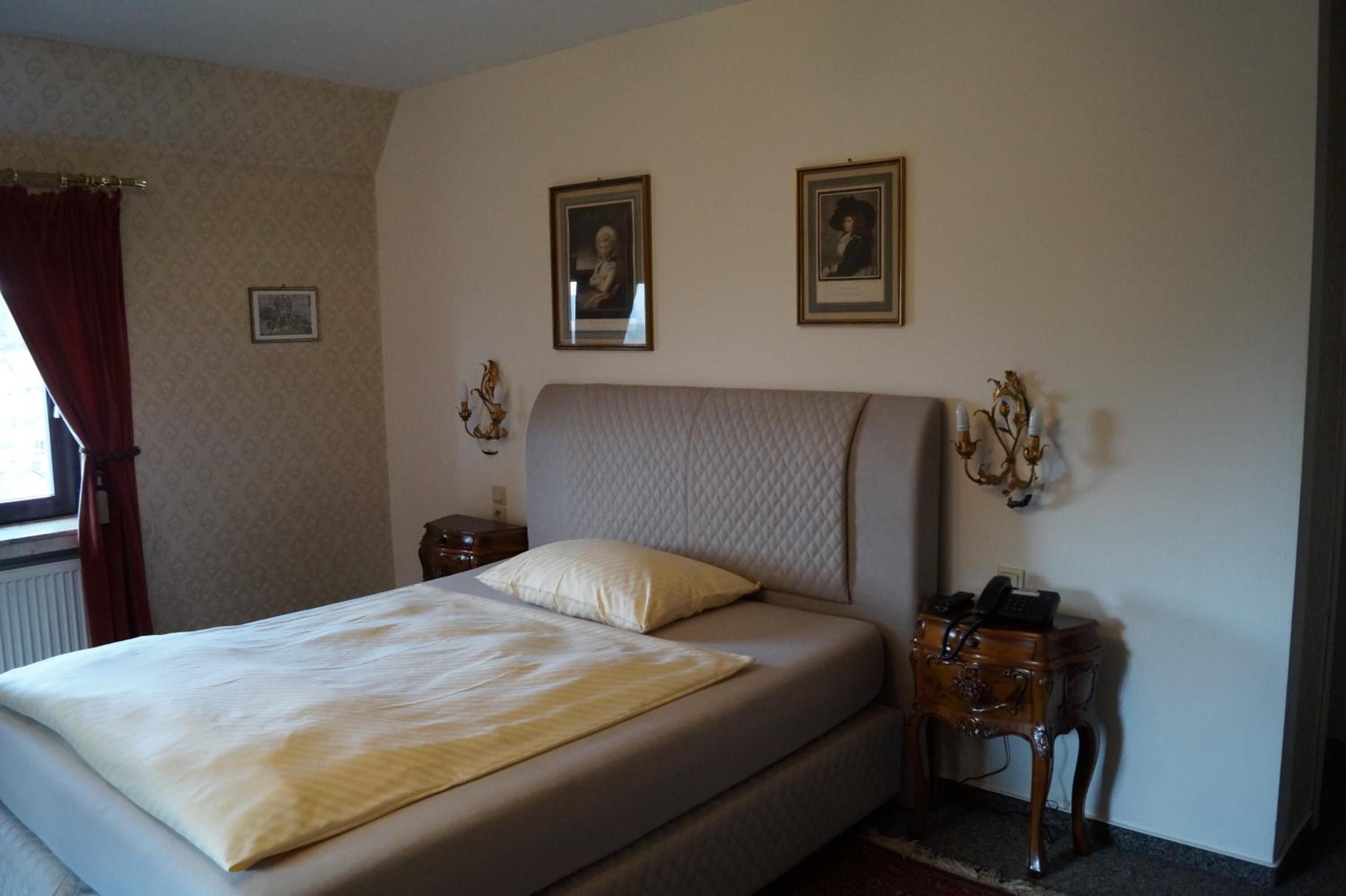 Ein Schlafzimmer mit einem Bett, zwei gerahmten Porträts an der Wand, einem Nachttisch mit Lampe und einem Fenster mit Vorhängen.