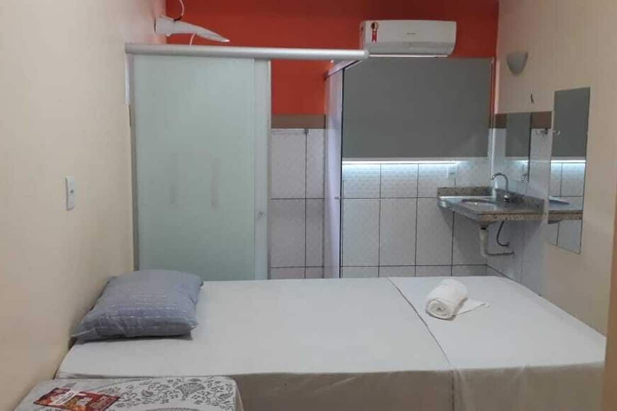A small, single-bed room with a bed, a pillow, a towel, a small table with a magazine, a sink, and a mirror.