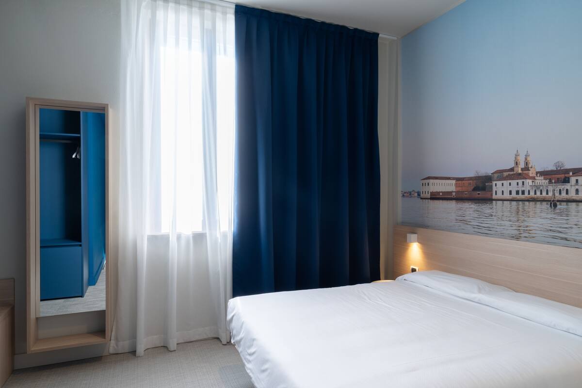 A hotel room with a bed, a wardrobe, a window with curtains, and a wall mural of a cityscape.