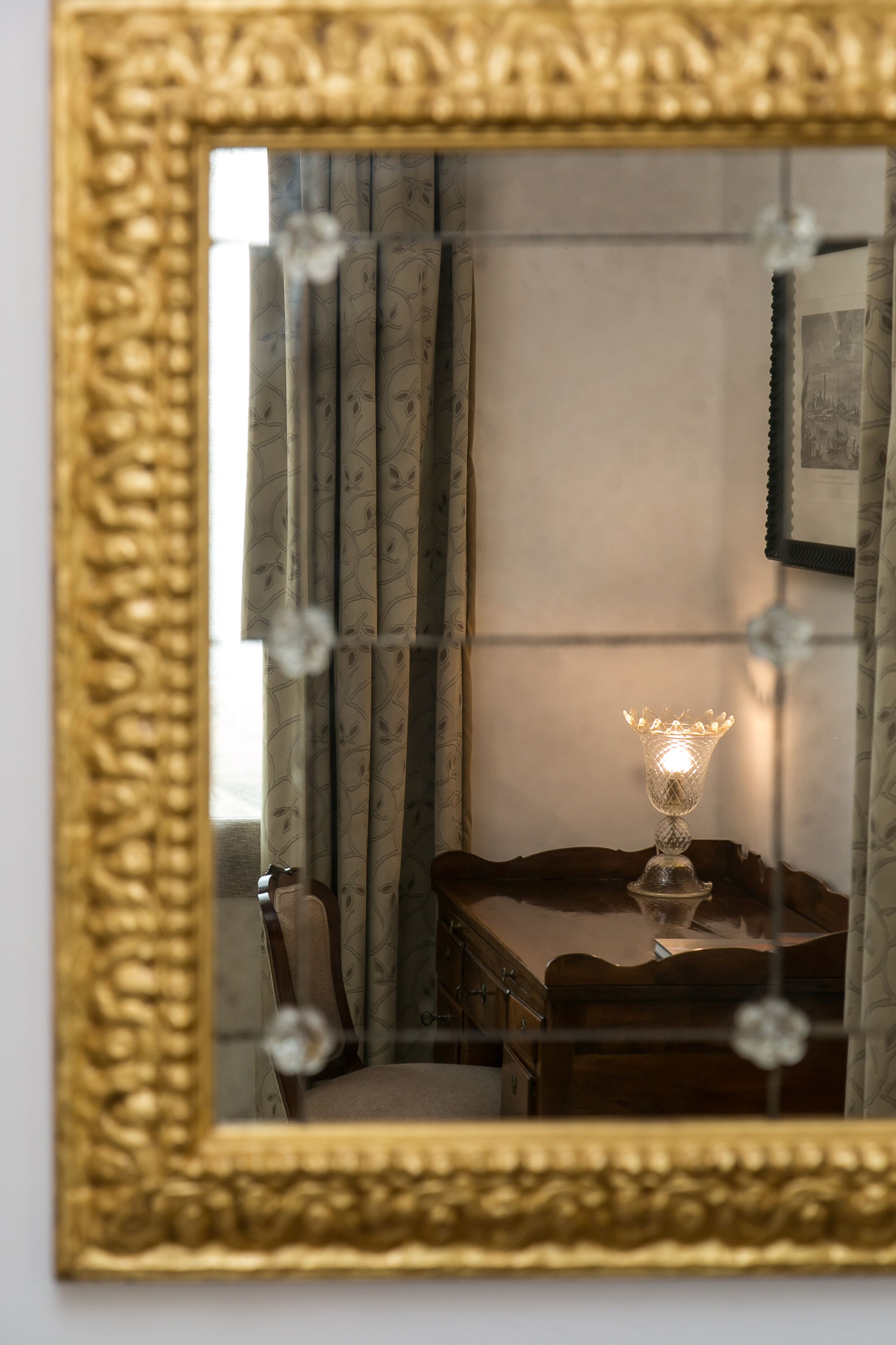 Un espejo con un marco dorado ornamentado refleja una habitación con una mesa, cortinas y un cuadro colgado en la pared.