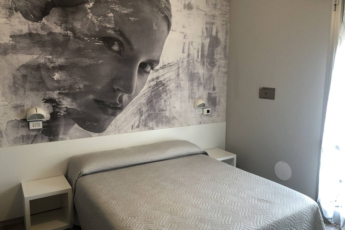 Une chambre à coucher comprenant un lit, une table de chevet, une lampe fixée au mur et un grand tableau abstrait.