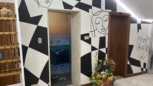 A hallway with geometric wall art, a wooden key rack, a door with a 'W' logo, and a floral arrangement. - Hotel Wega (Bellaria-Igea Marina)