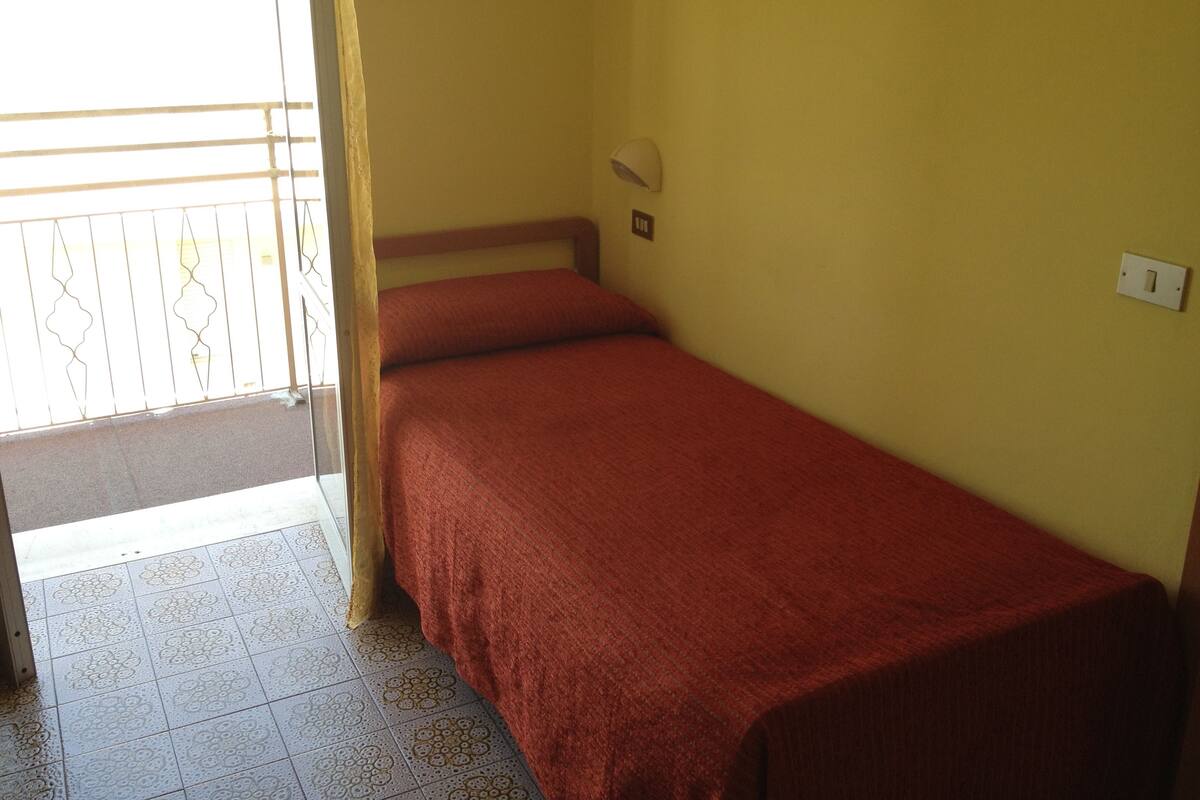 Une petite chambre avec un lit simple, un mur jaune uni, une fenêtre avec une grille en métal et une porte donnant sur un balcon extérieur.