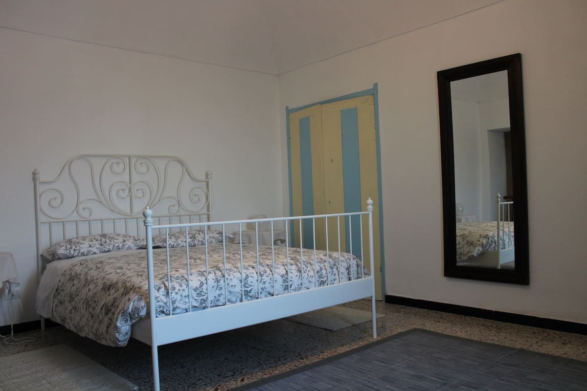 Una cama individual con estructura metálica y ropa de cama floral, un espejo en la pared y una puerta con franjas azules y amarillas.