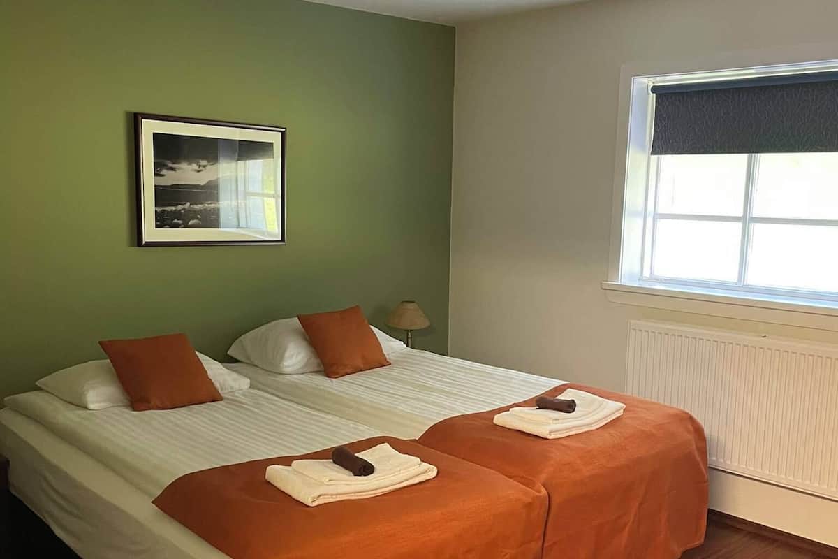 Een tweepersoonsbed met twee oranje dekens, een ingelijst schilderij aan de muur, een raam met een donkerkleurige zonwering en een radiator.