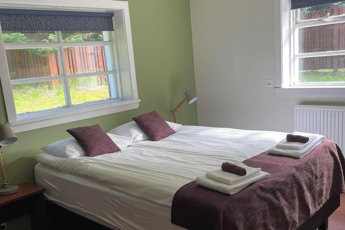 Een slaapkamer met een bed, nachtkastje, lamp en een raam met uitzicht op een houten schutting en groen.