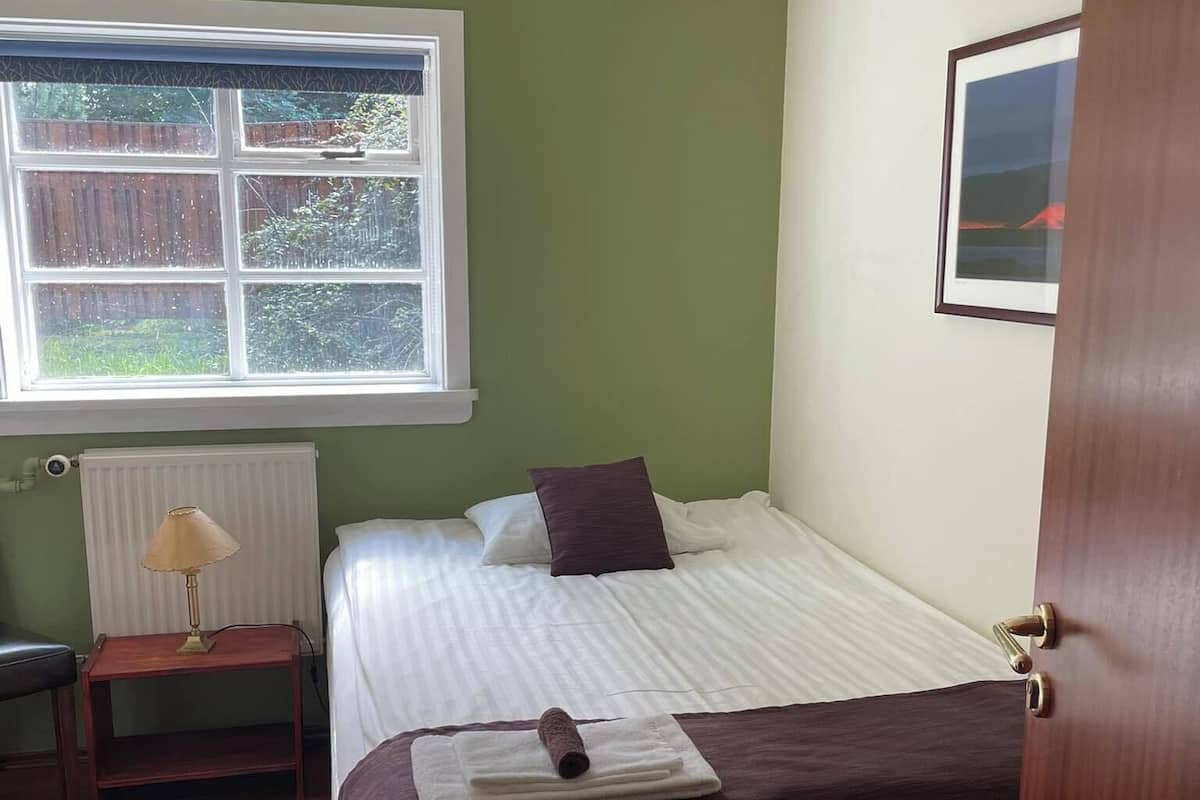 Een slaapkamer met een bed, nachtkastje, lamp en een raam met uitzicht op groen.