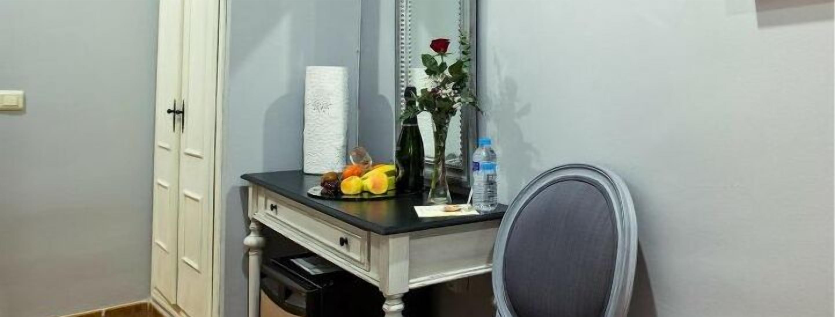 Un pequeño escritorio con un espejo, un florero con una rosa roja, una botella de agua y algunas frutas.