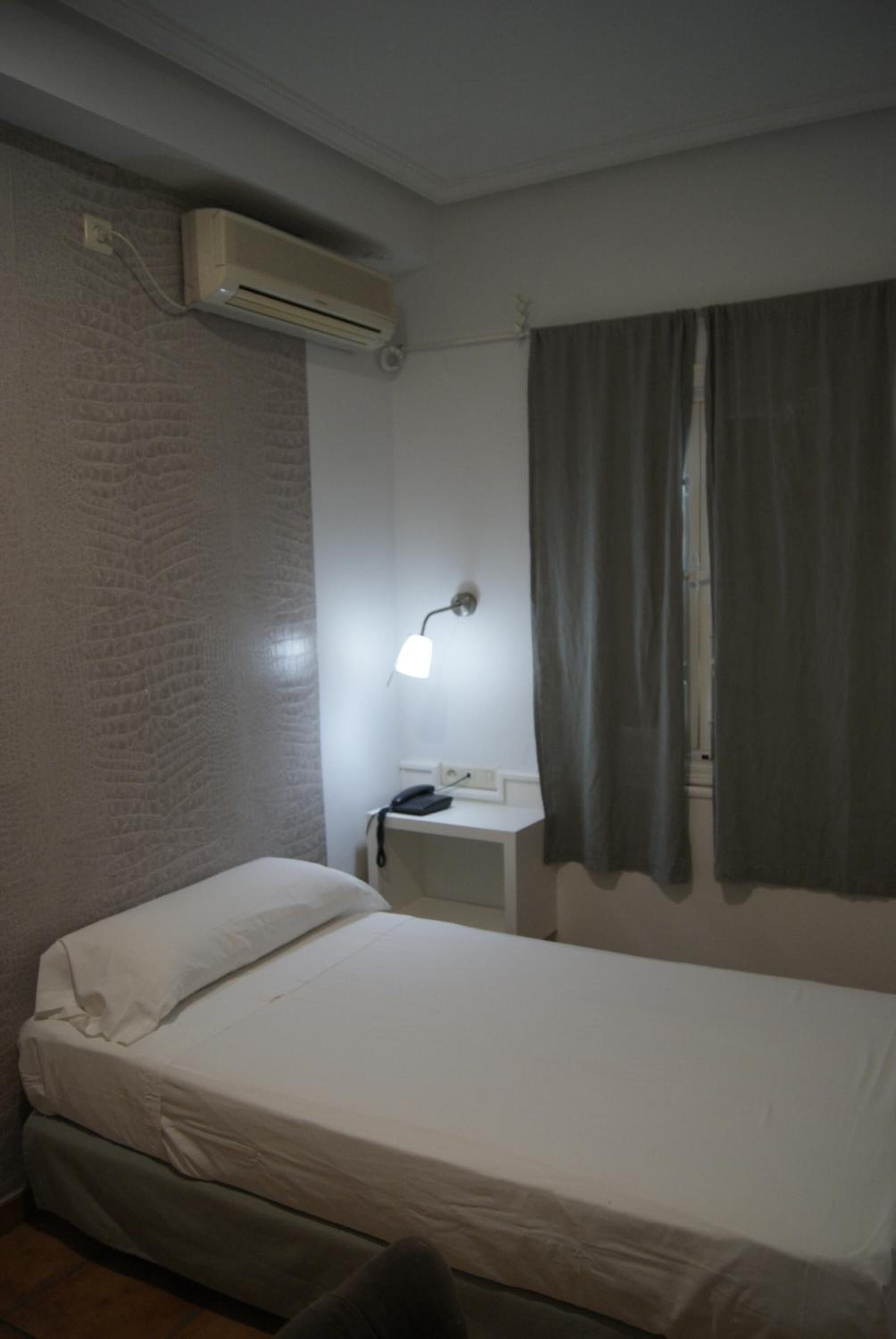A small, single-bed room with a white bed, a bedside table with a lamp, a wall-mounted air conditioner, and a window with curtains.