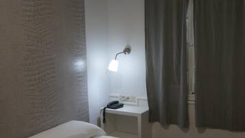 A small, single-bed room with a white bed, a bedside table with a lamp, a wall-mounted air conditioner, and a window with curtains.