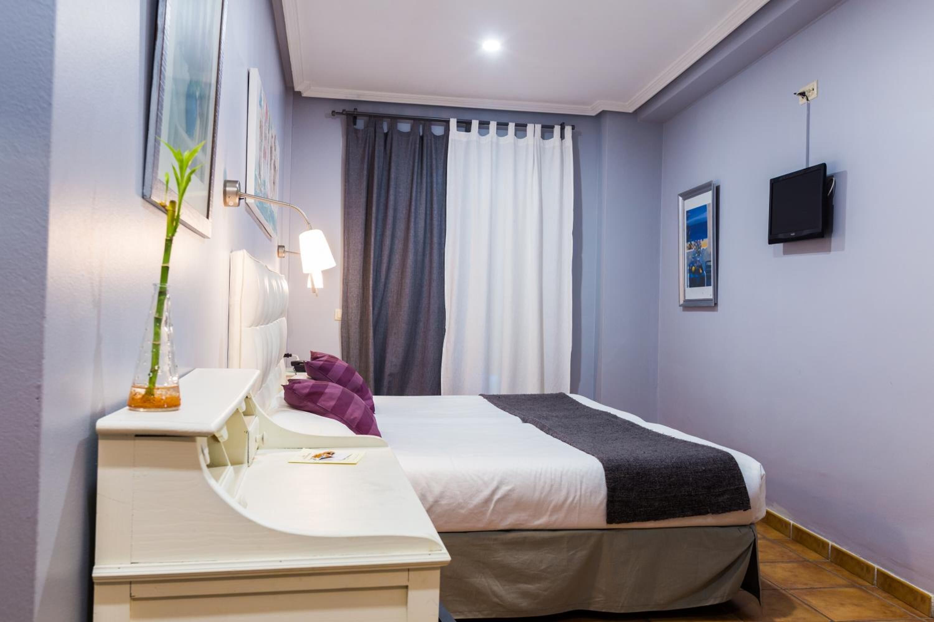 Habitación de hotel con una cama, un escritorio con lámpara, un televisor y una ventana con cortinas.