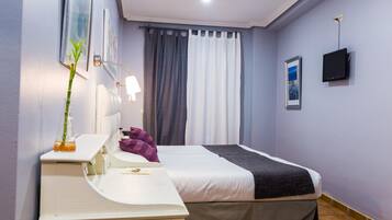 Habitación de hotel con una cama, un escritorio con lámpara, un televisor y una ventana con cortinas.