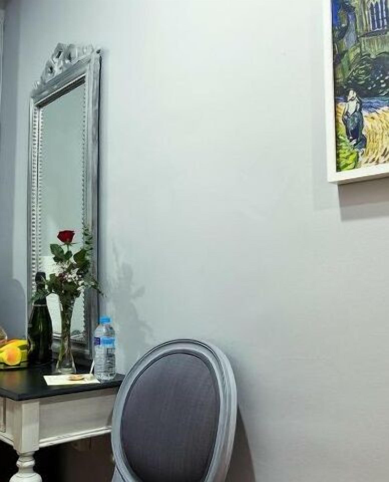 A room with a large mirror, a painting on the wall, a chair, a table with a vase of flowers, and a water bottle.