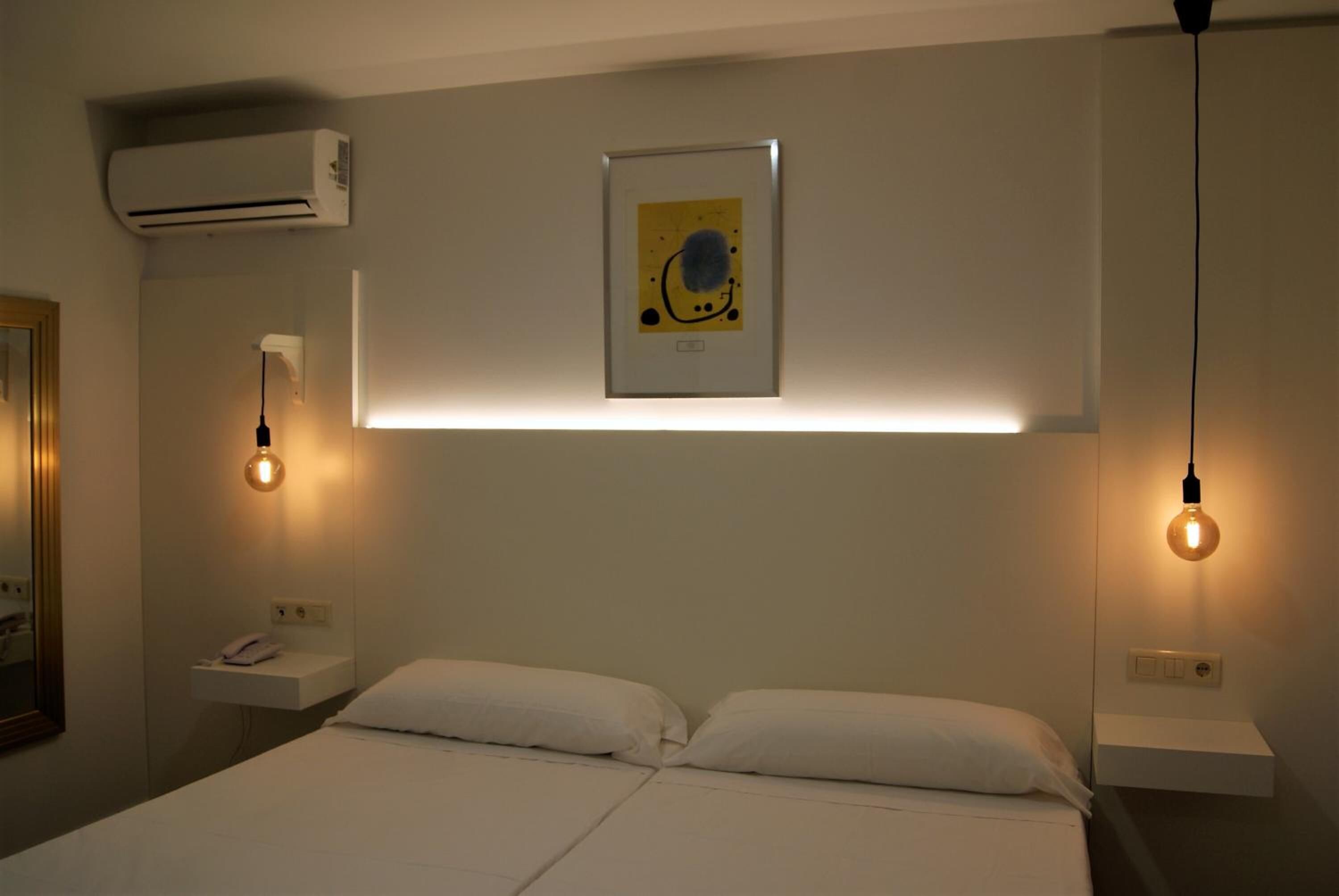 A hotel room with a bed, bedside tables, an air conditioner, and a framed artwork on the wall.