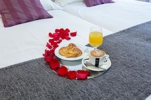 A breakfast tray with croissant, coffee, and orange juice on a bed with a grey blanket and red rose petals. - Hotel La Encomienda (Moraleja)