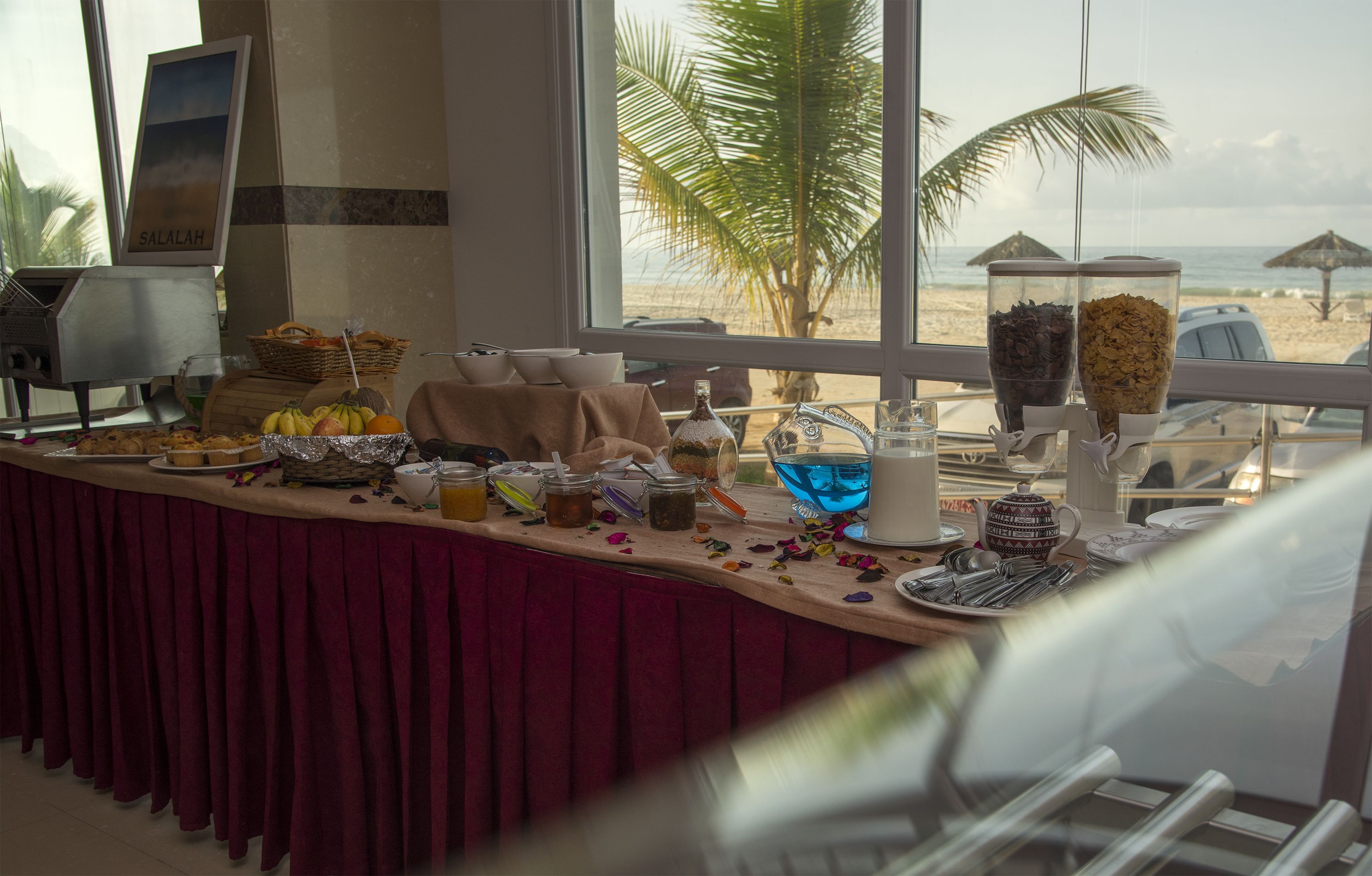 Una mesa de buffet con diversos alimentos y condimentos, una botella de salsa y vistas a la playa a través de amplias ventanas.