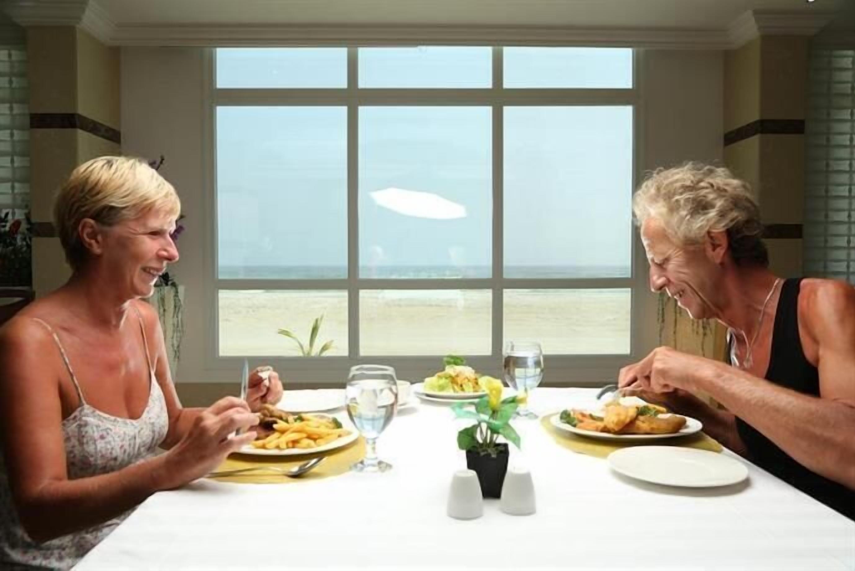 Duas pessoas jantando em uma mesa com vista para o mar através de grandes janelas.