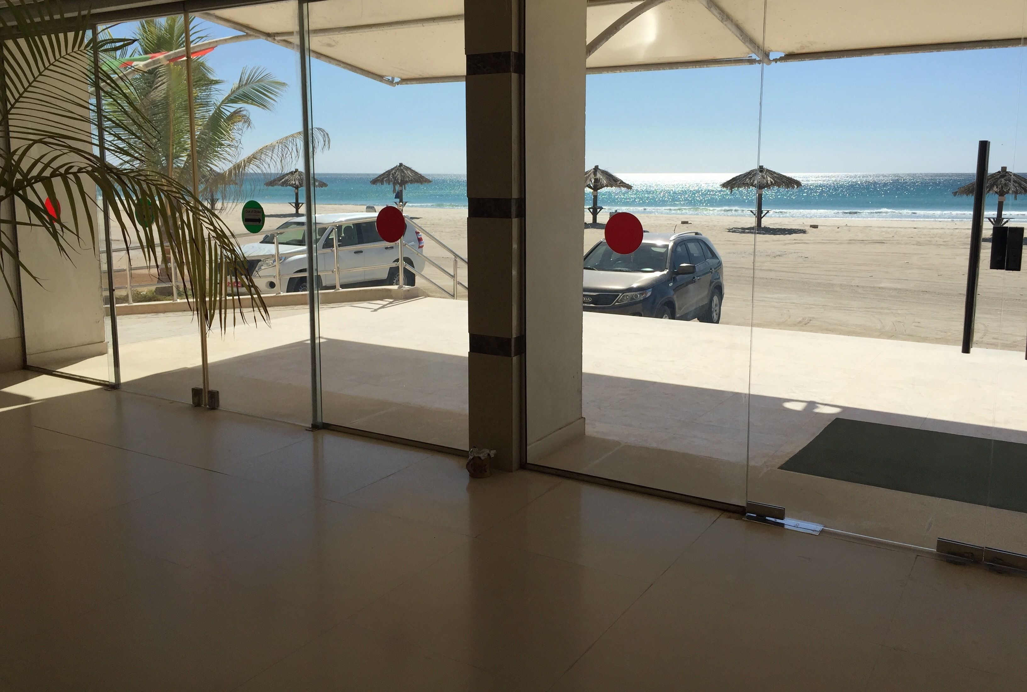 A beach view from a covered area with palm trees, umbrellas, and parked cars.