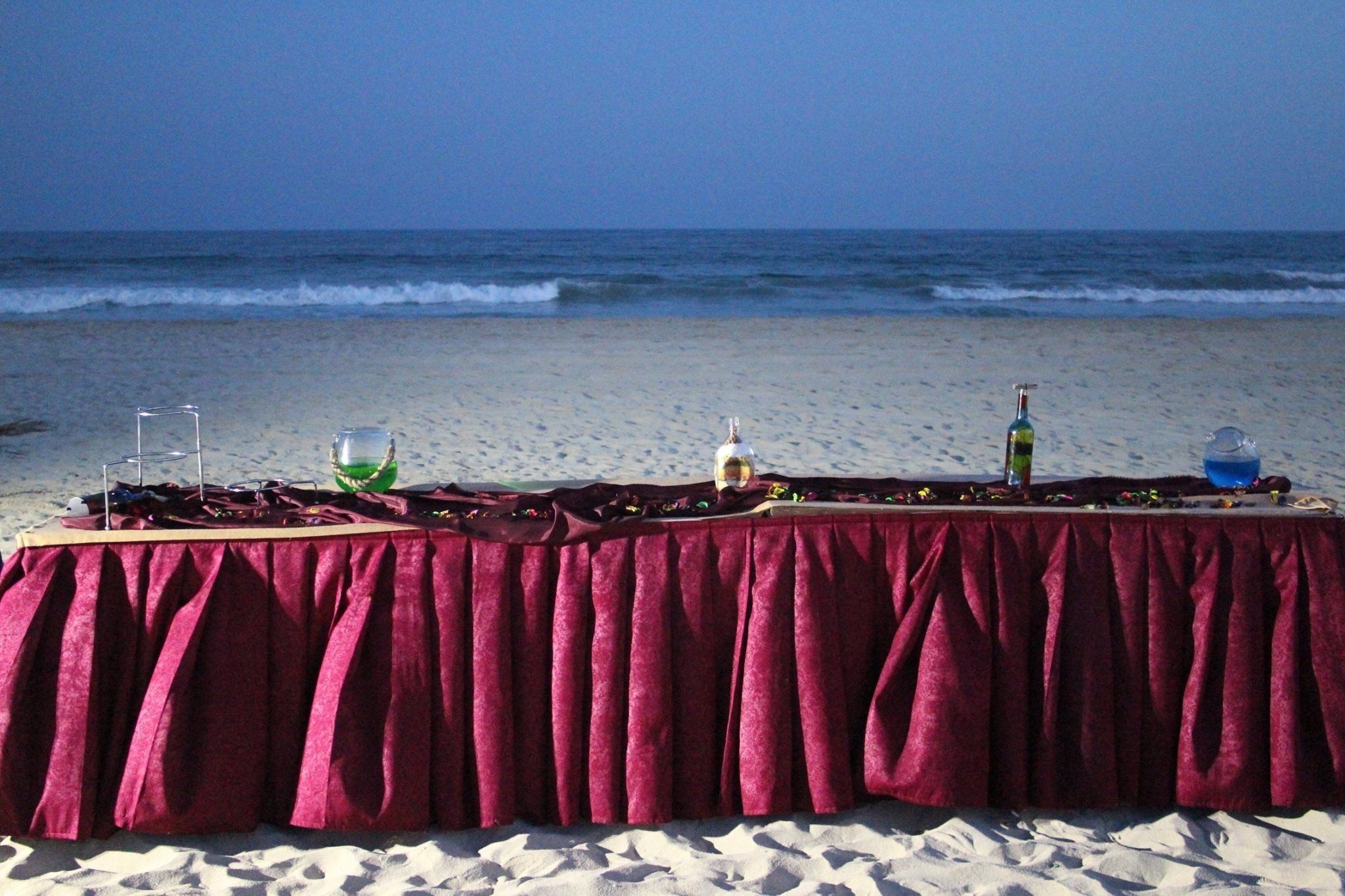 A table set up on a beach with a red tablecloth, bottles, and glasses.