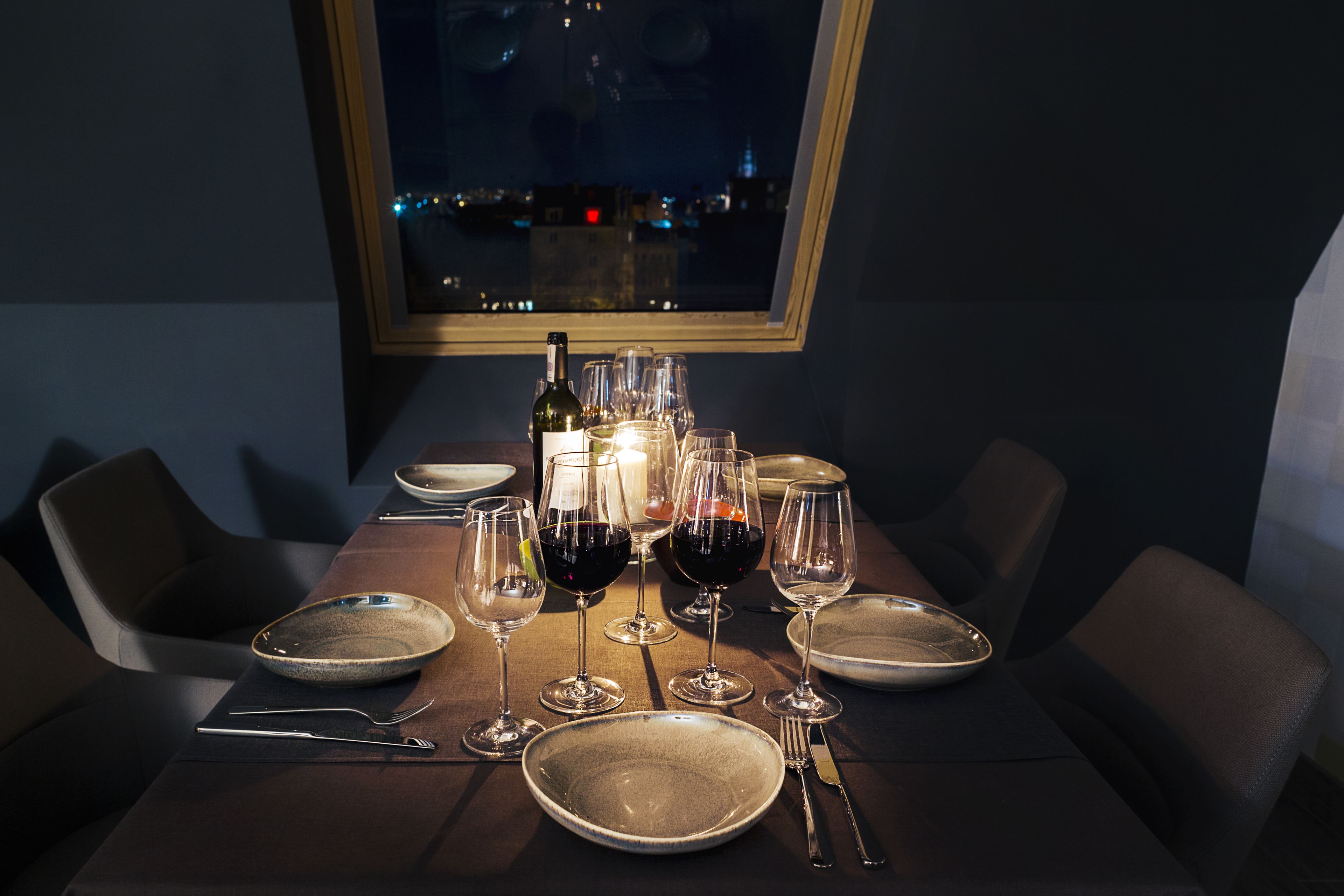 Tavola imbandita per sei persone, con calici da vino, piatti, posate, una candela e vista sulla città attraverso una finestra.