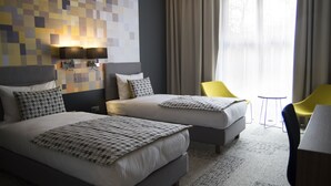 A hotel room with two beds, a small table, a chair, and a large window with curtains. - B&B HOTEL Gdańsk Old Town (Gdansk)