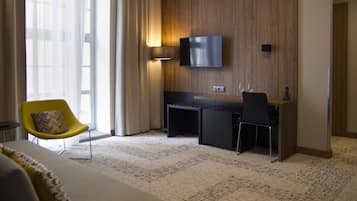 A modern hotel room with a large window, a flat-screen TV mounted on the wall, a desk with a chair, and a yellow armchair.