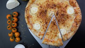 Pizza mit einem fehlenden Stück, umgeben von Tomaten, einer Olivenölflasche und einem Salzstreuer auf dunklem Untergrund.