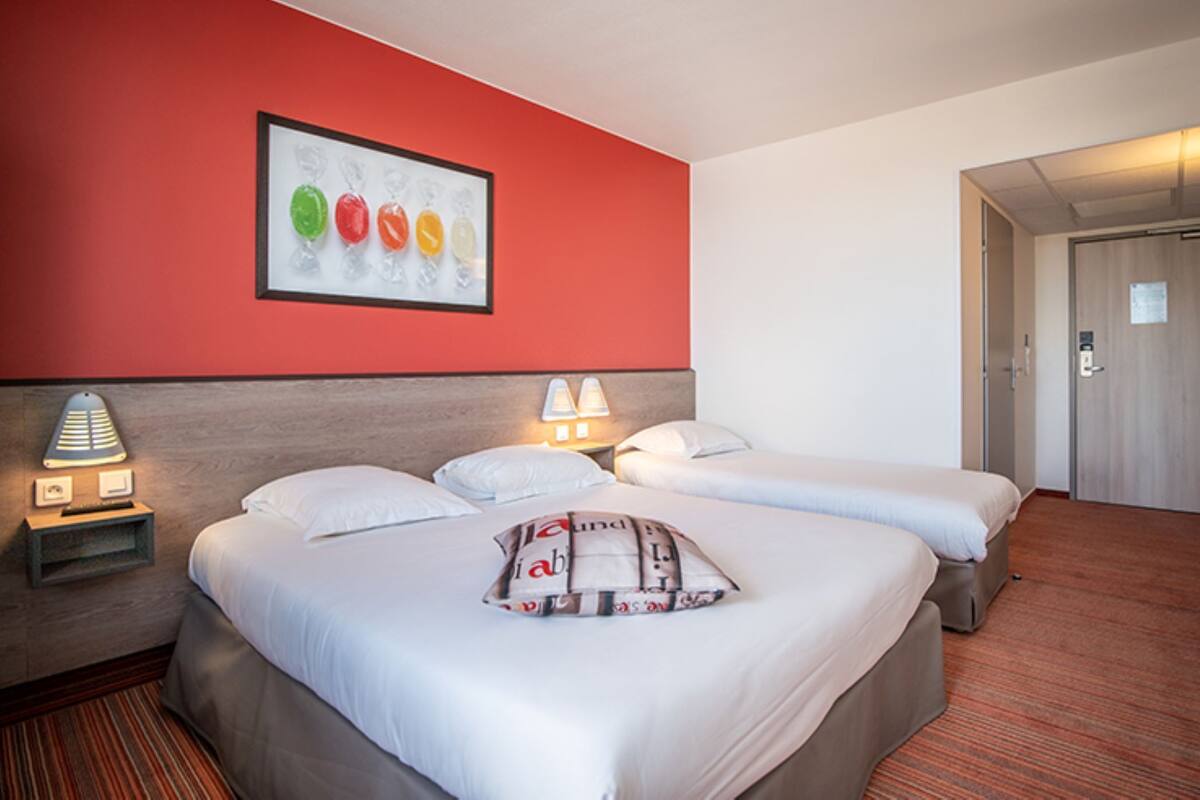 A hotel room with three beds, a red accent wall, a framed artwork, and a door leading to a hallway.