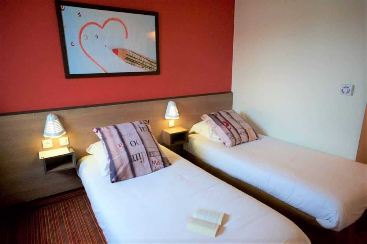A hotel room with two single beds, each with a bedside lamp and a pillow with text. There is a framed picture on the wall above the beds.