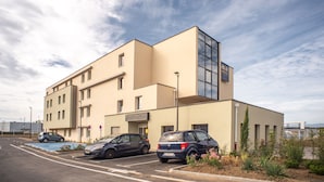 A modern hotel with a flat facade, multiple windows, and a parking lot with several cars. - Ace hôtel Annemasse Genève (Ville-la-Grand)